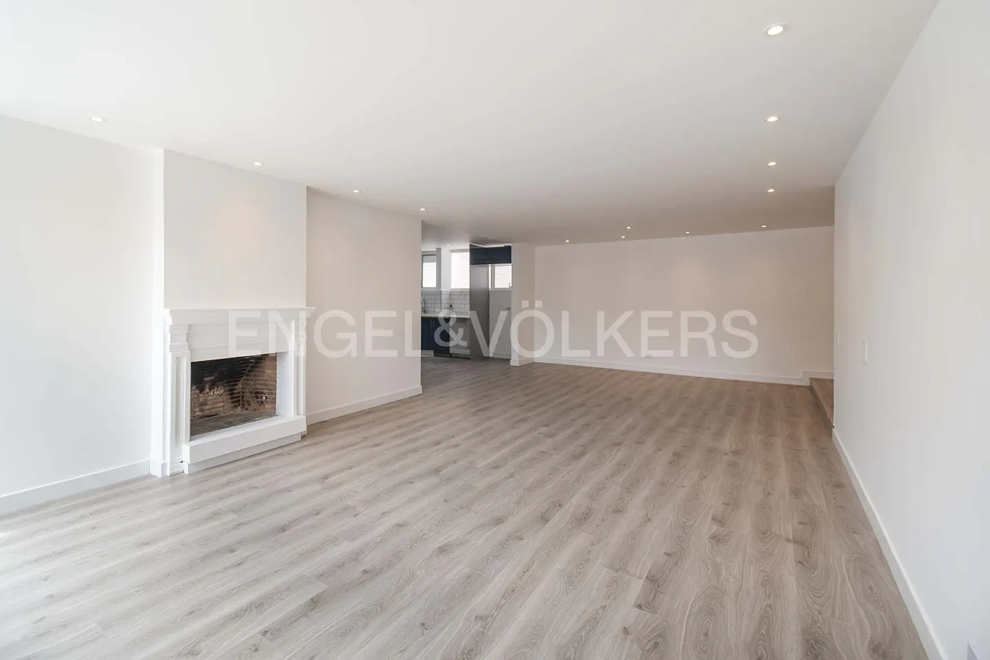 Bright, empty living room with light wood floors, white walls, and a fireplace. A kitchen is visible in the background.