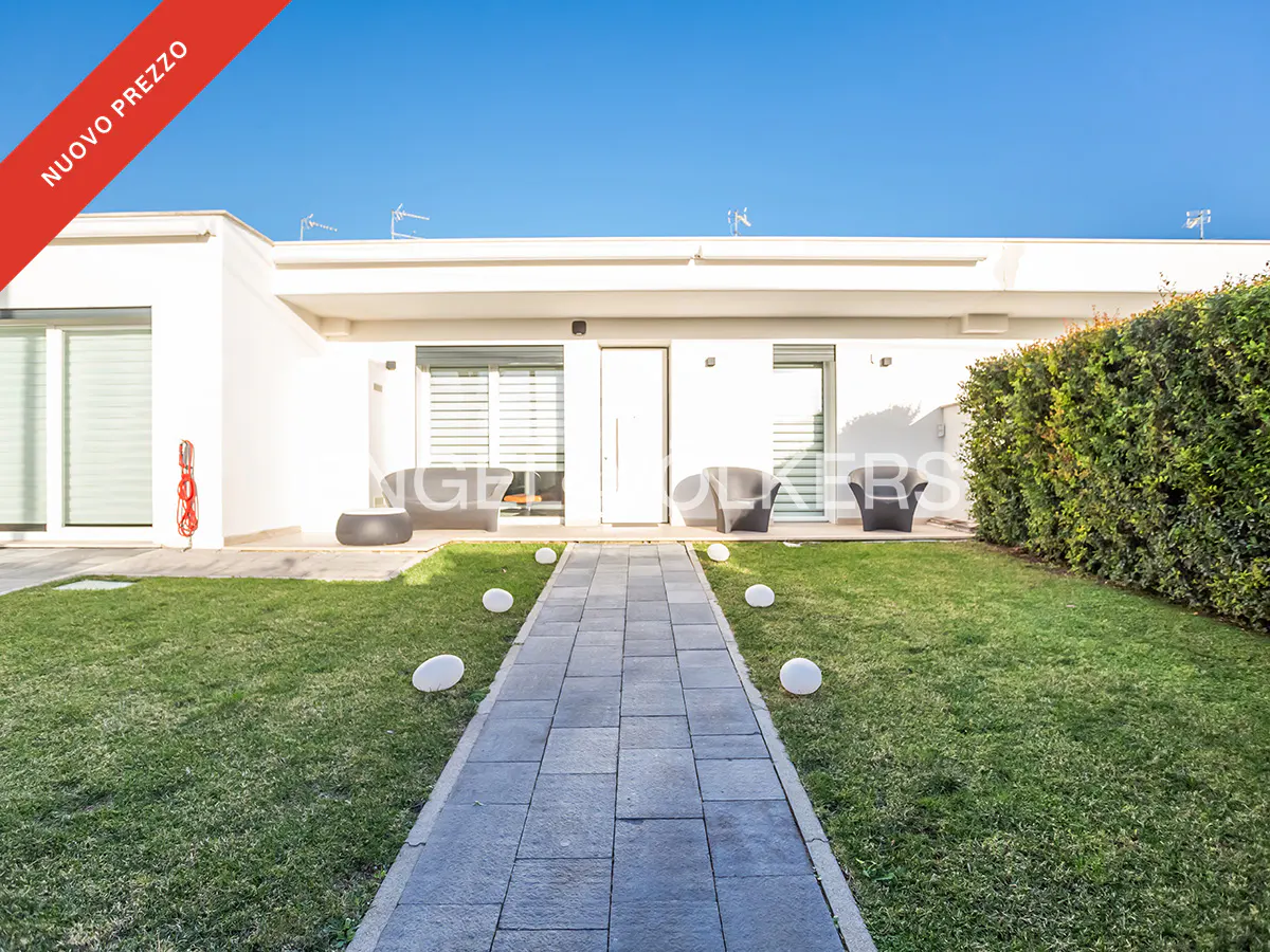 Modern white house with a stone walkway, green lawn, and outdoor seating under a clear blue sky. "Nuovo Prezzo" red banner.