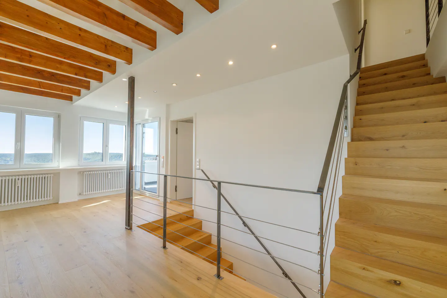 Bright, modern home interior with wood floors, exposed beams, and staircase. Large windows offer a scenic view. Metal railings add a contemporary touch.