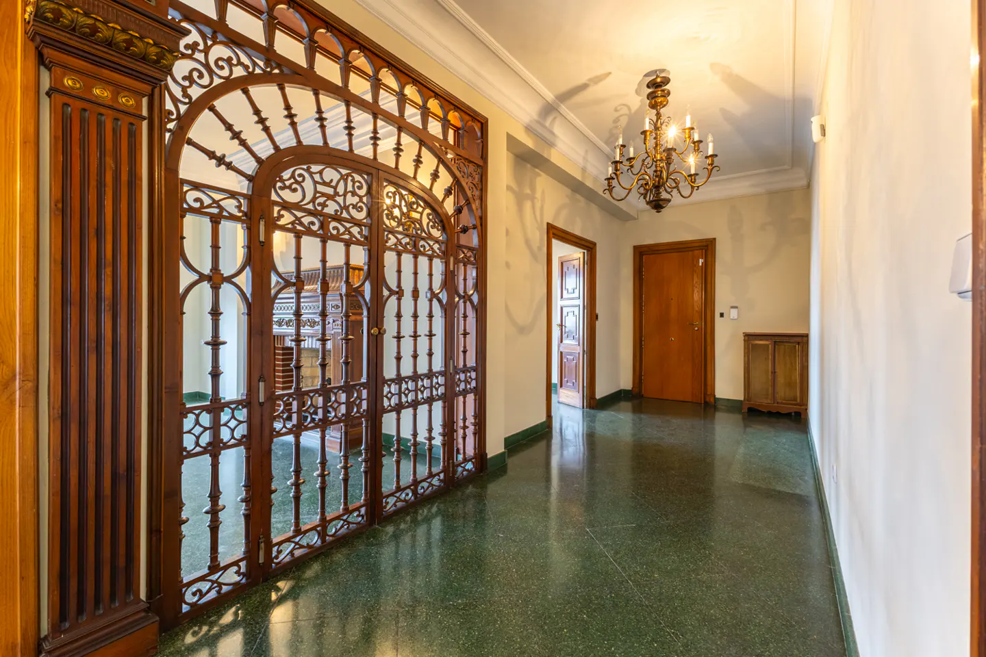 Hallway with green terrazzo floor, ornate wooden gate, chandelier, and wooden doors leading to other rooms.
