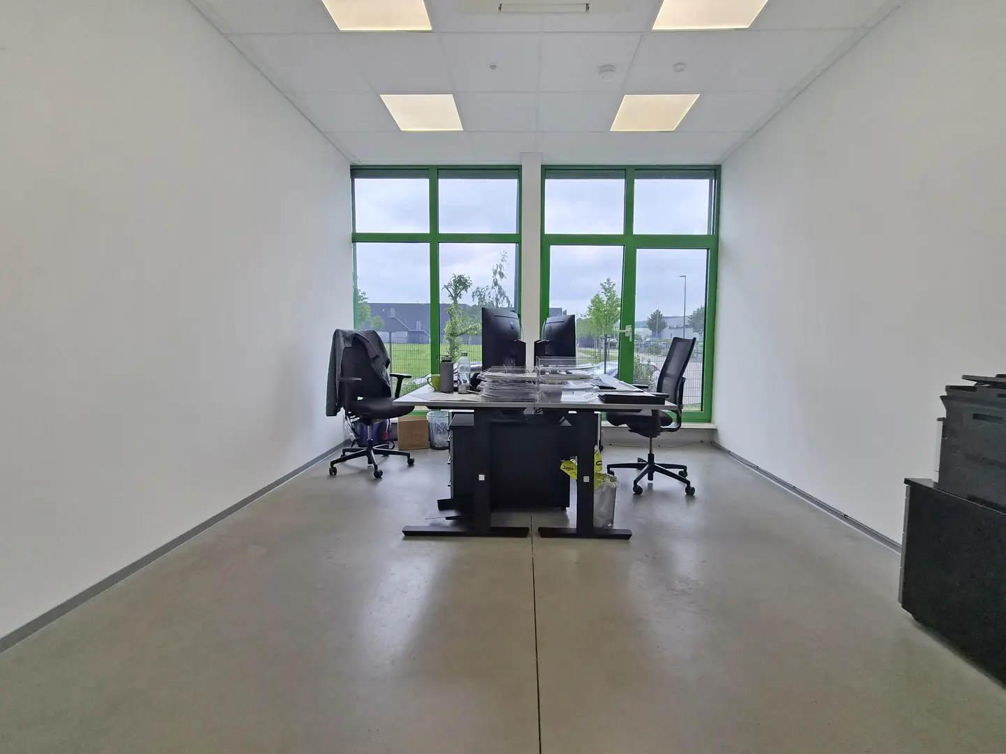 Bright office space with white walls, a concrete floor, and green-framed windows. Desks, chairs, and computer monitors are visible.