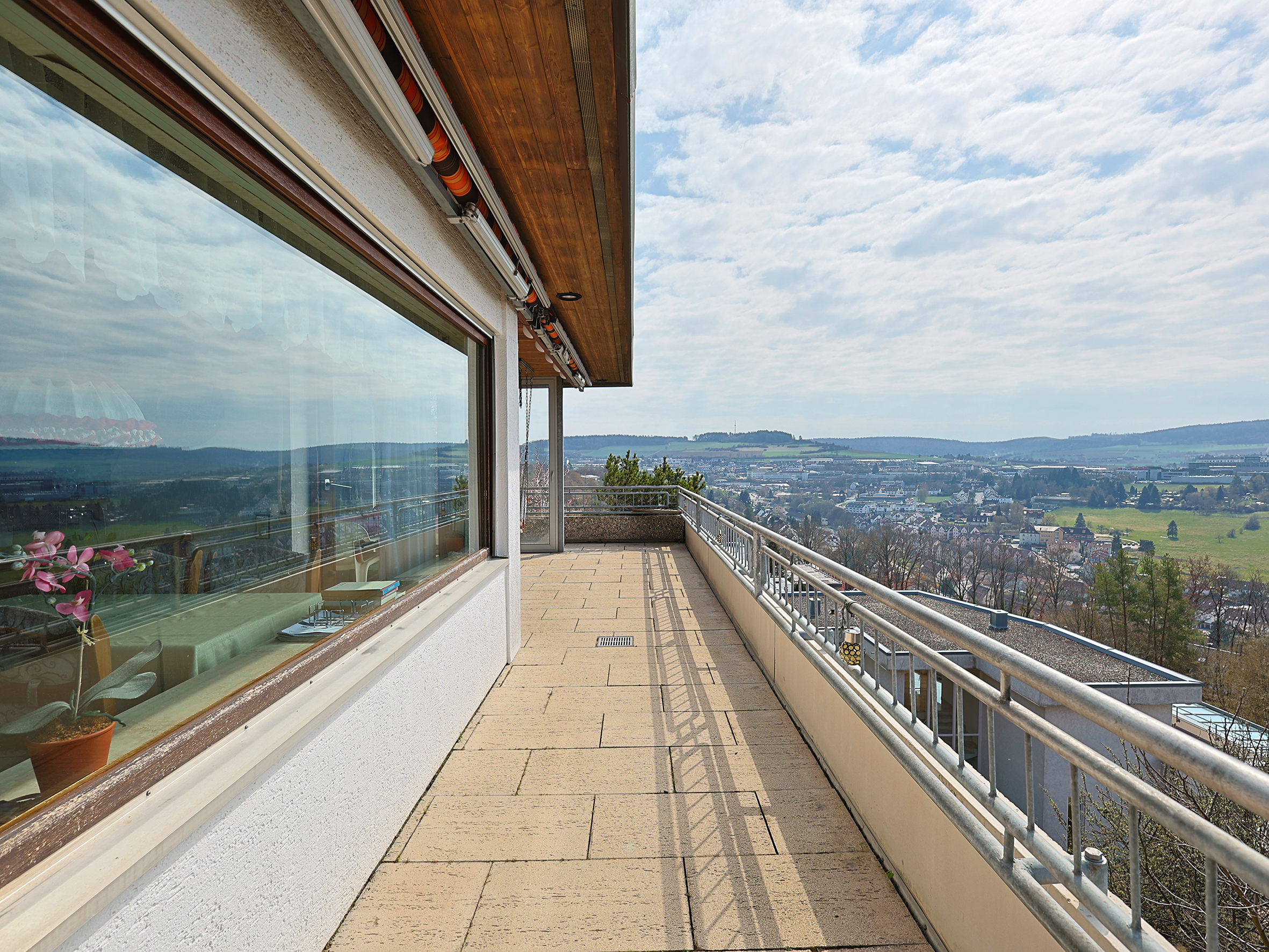 A long, stone balcony with a metal railing overlooks a city and green hills under a cloudy sky. A large window reflects the view.