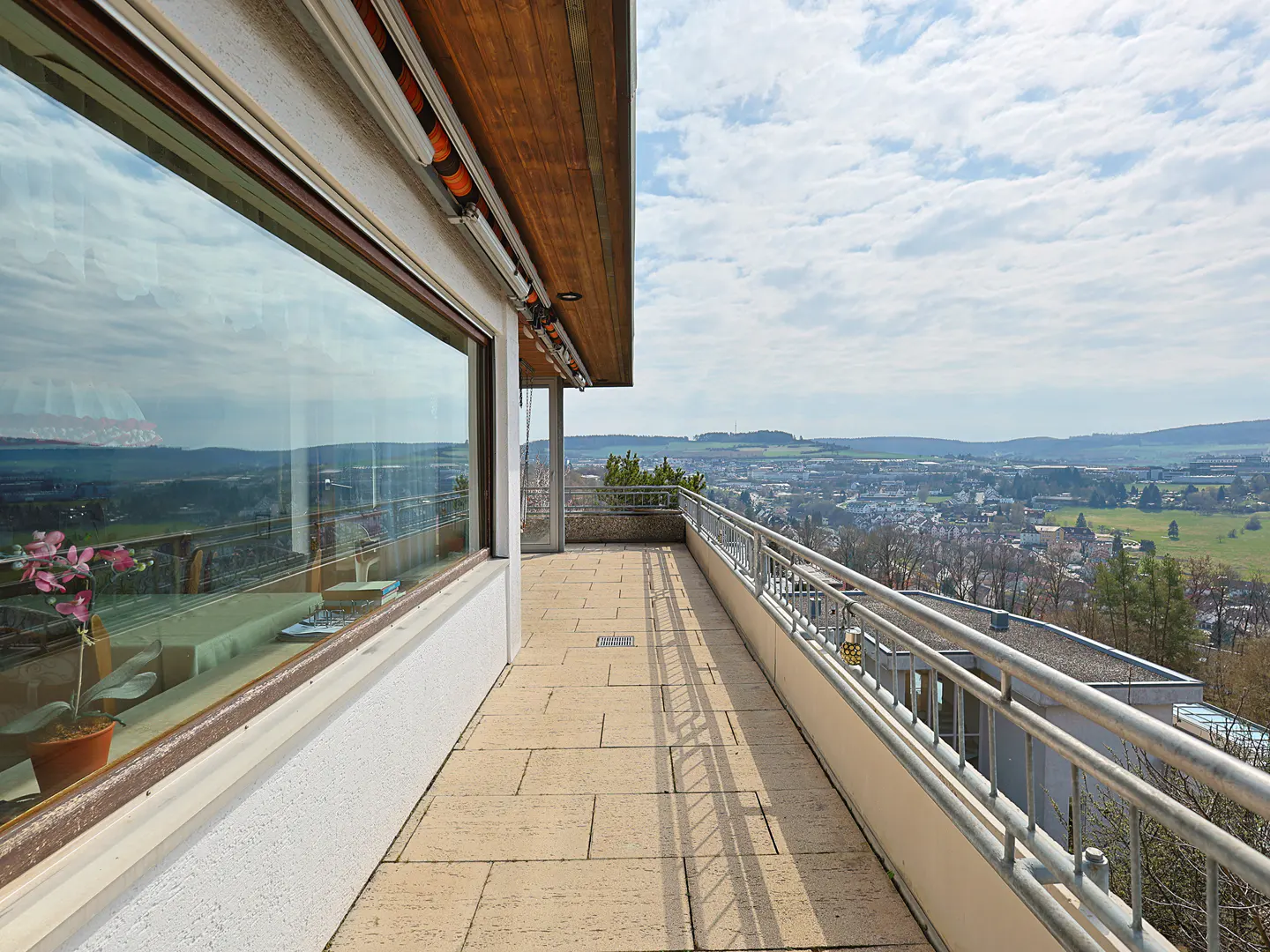 A long, stone balcony with a metal railing overlooks a city and green hills under a cloudy sky. A large window reflects the view.