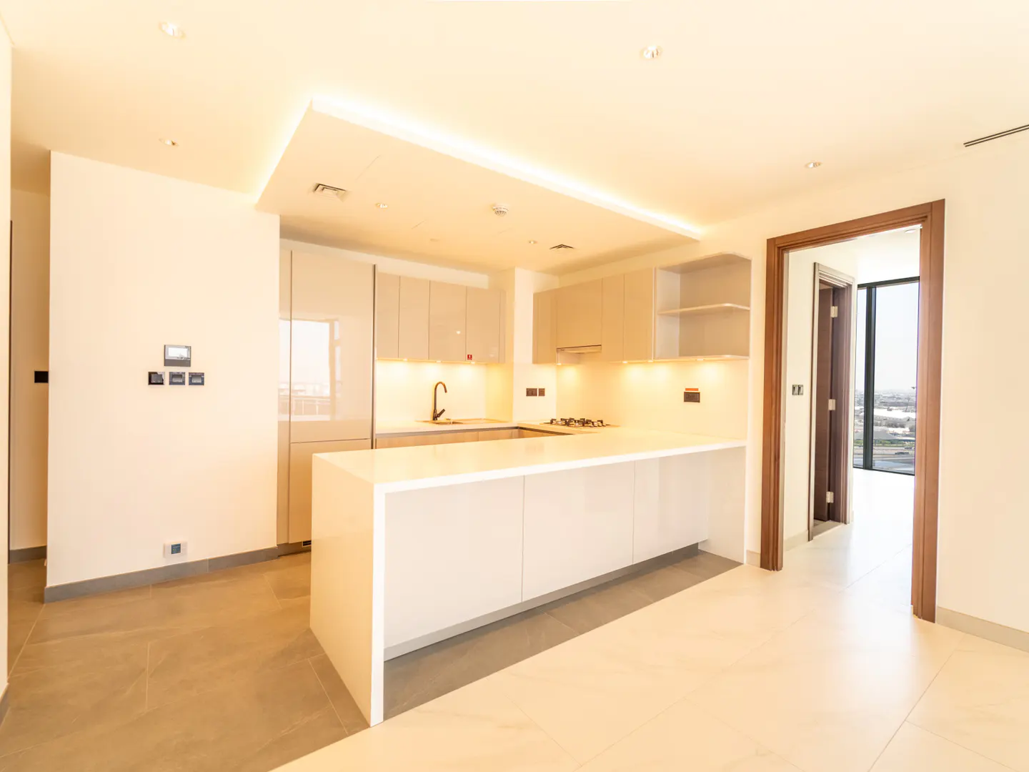 Modern kitchen with white island, beige cabinets, under-cabinet lighting, and a doorway leading to a bright room.