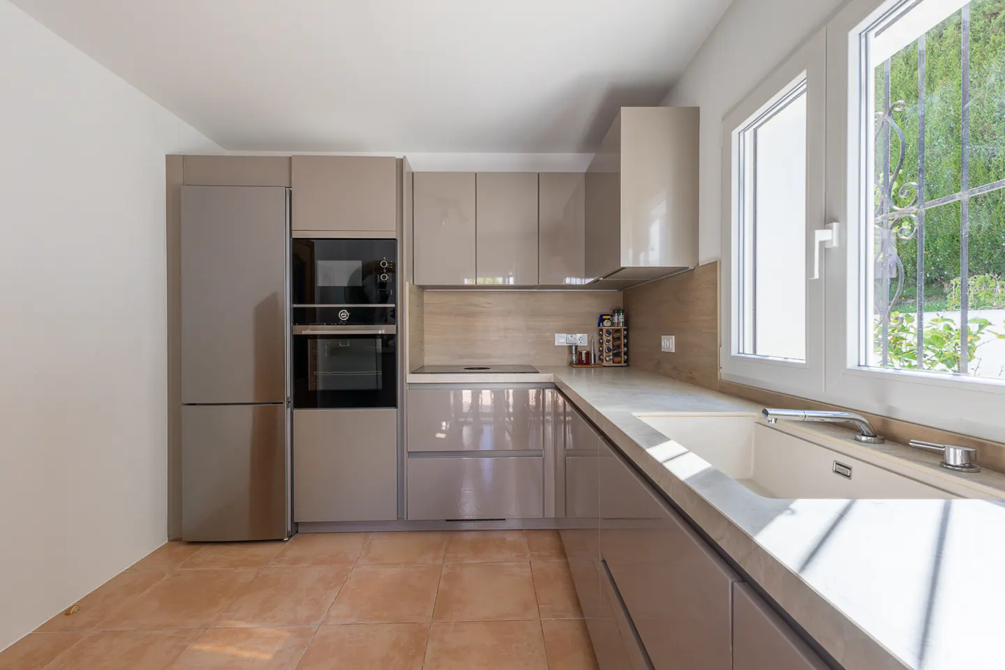 Modern kitchen with beige cabinets, stainless steel appliances, and a large window overlooking greenery. Brown tile floor.