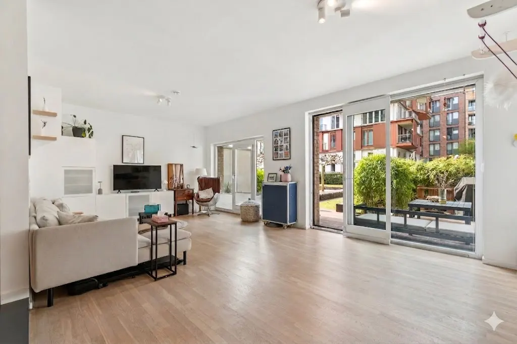 Bright living room with hardwood floors, white walls, and sliding glass doors to a patio with greenery and a view of a brick building.