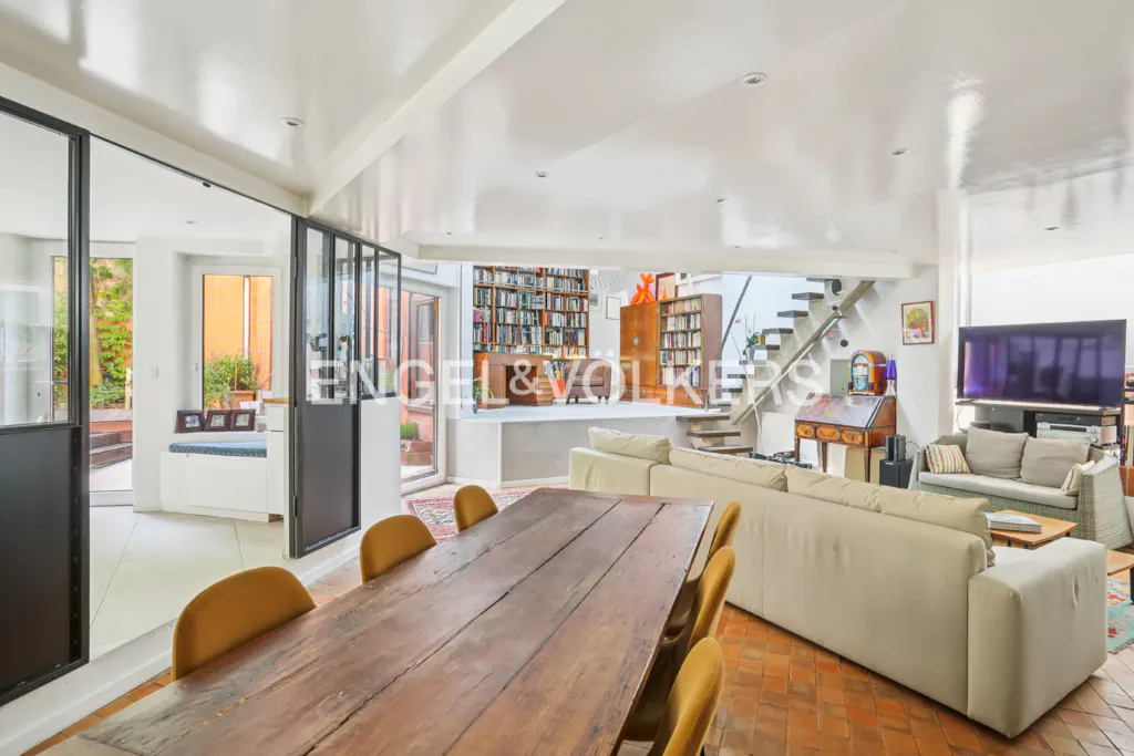 Open-concept living space with a long wooden table, yellow chairs, and a cream sofa. A staircase leads to a library with floor-to-ceiling bookshelves.