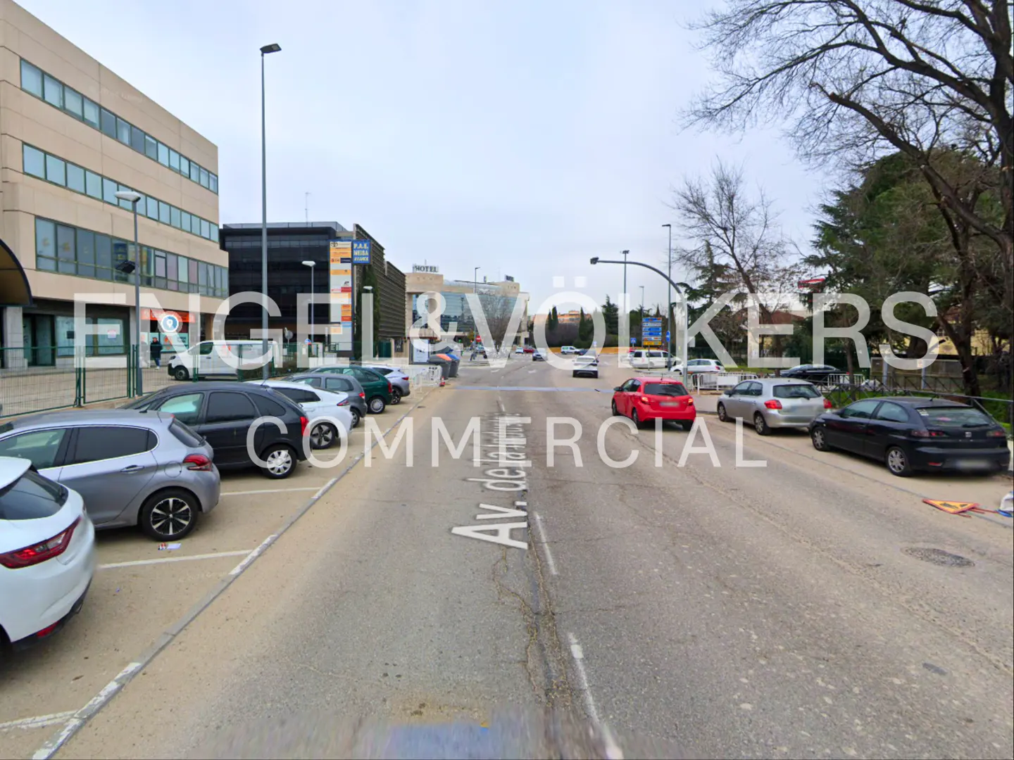 Street view of Av. de la Victoria with parked cars, buildings, and trees under a cloudy sky. A red car is driving on the road.