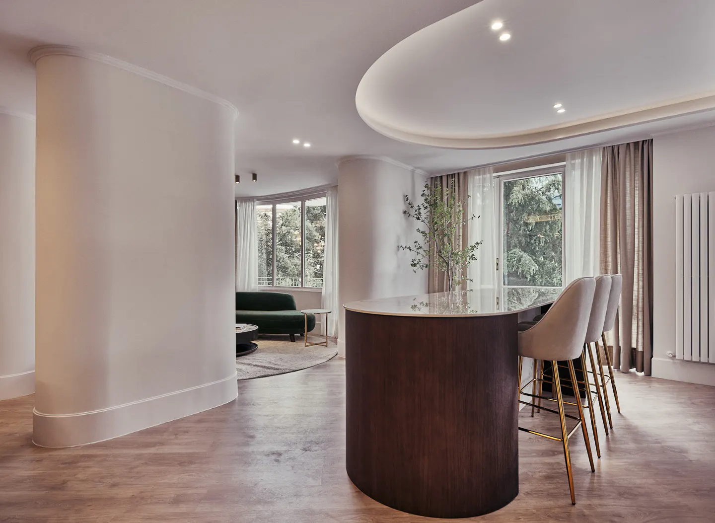 Open-concept living space with curved walls, wood floors, and a dark wood island with three beige chairs. A green sofa is visible in the background.