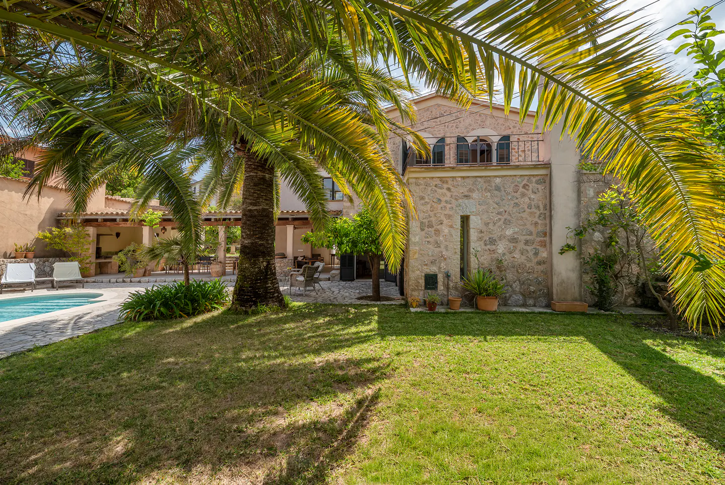 Exterior view of a stone house with a green lawn, palm trees, and a swimming pool in the background.