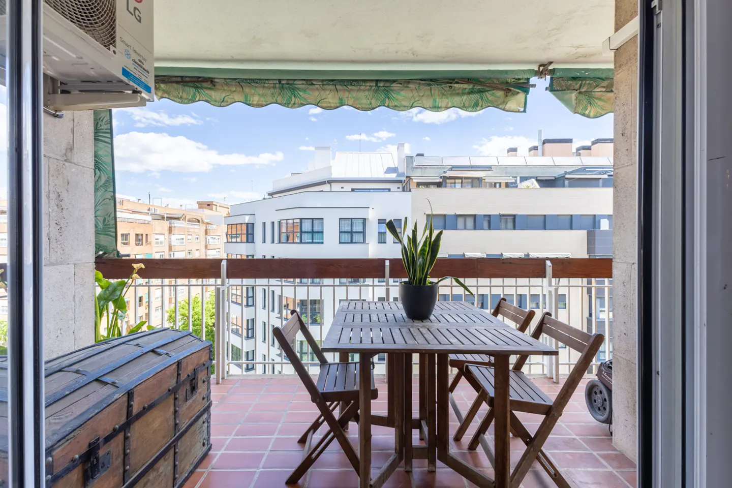 Balcony with a wooden table, four chairs, and a potted plant. A chest sits to the side, with city buildings in the background.