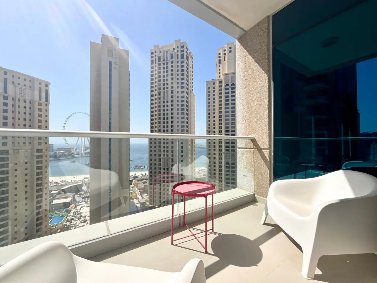 Balcony view with white chairs, red table, and city skyline. Ocean and Ferris wheel visible in the background.