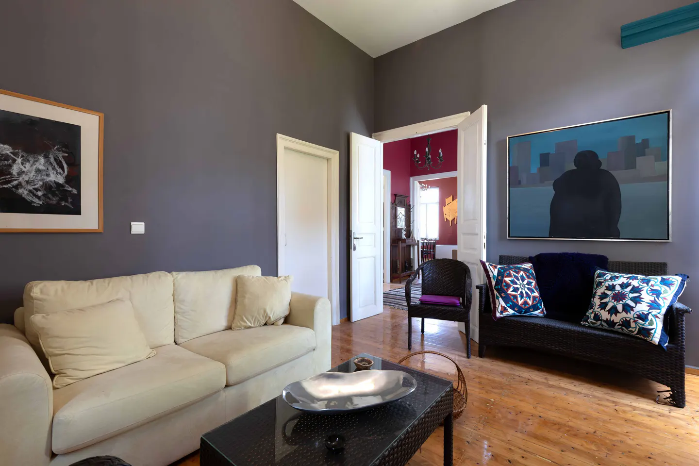 Living room with gray walls, light wood floors, and art. A cream sofa and black wicker furniture are arranged around a black coffee table.