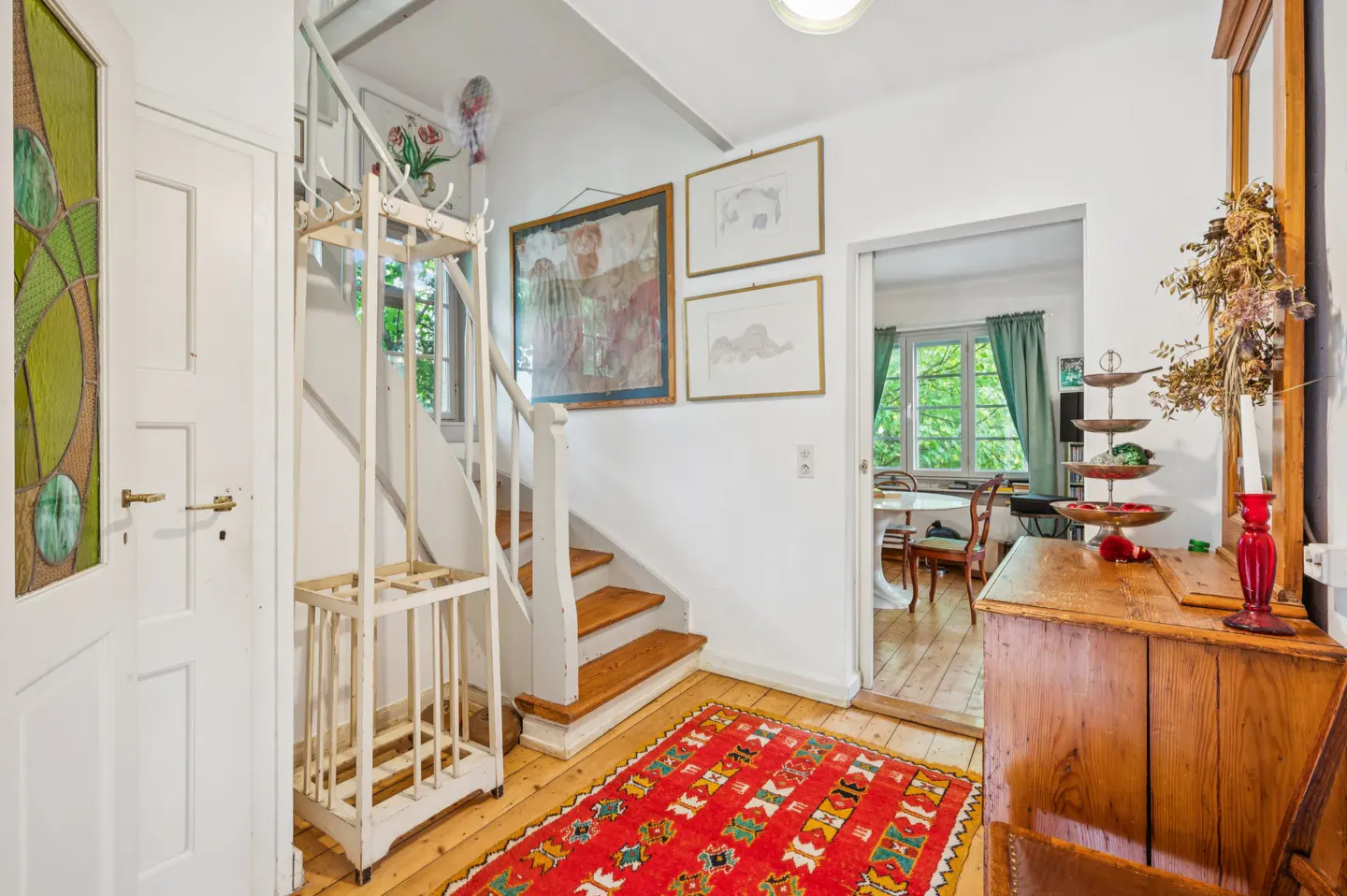 Entryway with white walls, wooden floors, and a red patterned rug. A staircase leads up, and a doorway shows a dining room. Art adorns the walls.