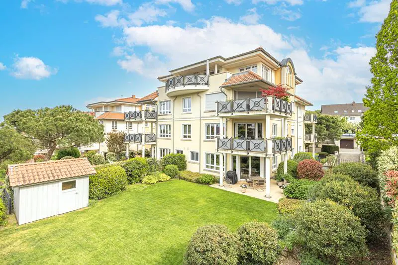 Three-story apartment building with balconies, a green lawn, and a white shed under a blue sky.