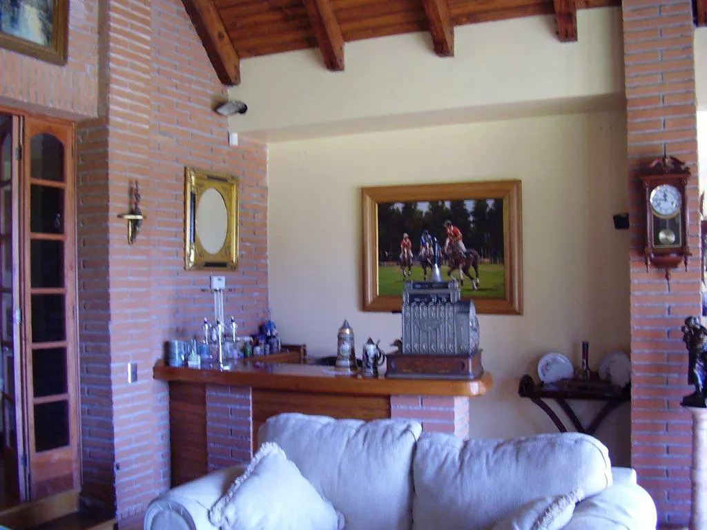 Living room with brick walls, wood ceiling, and cream sofa. A painting of polo players hangs above a bar with glassware and an antique cash register.