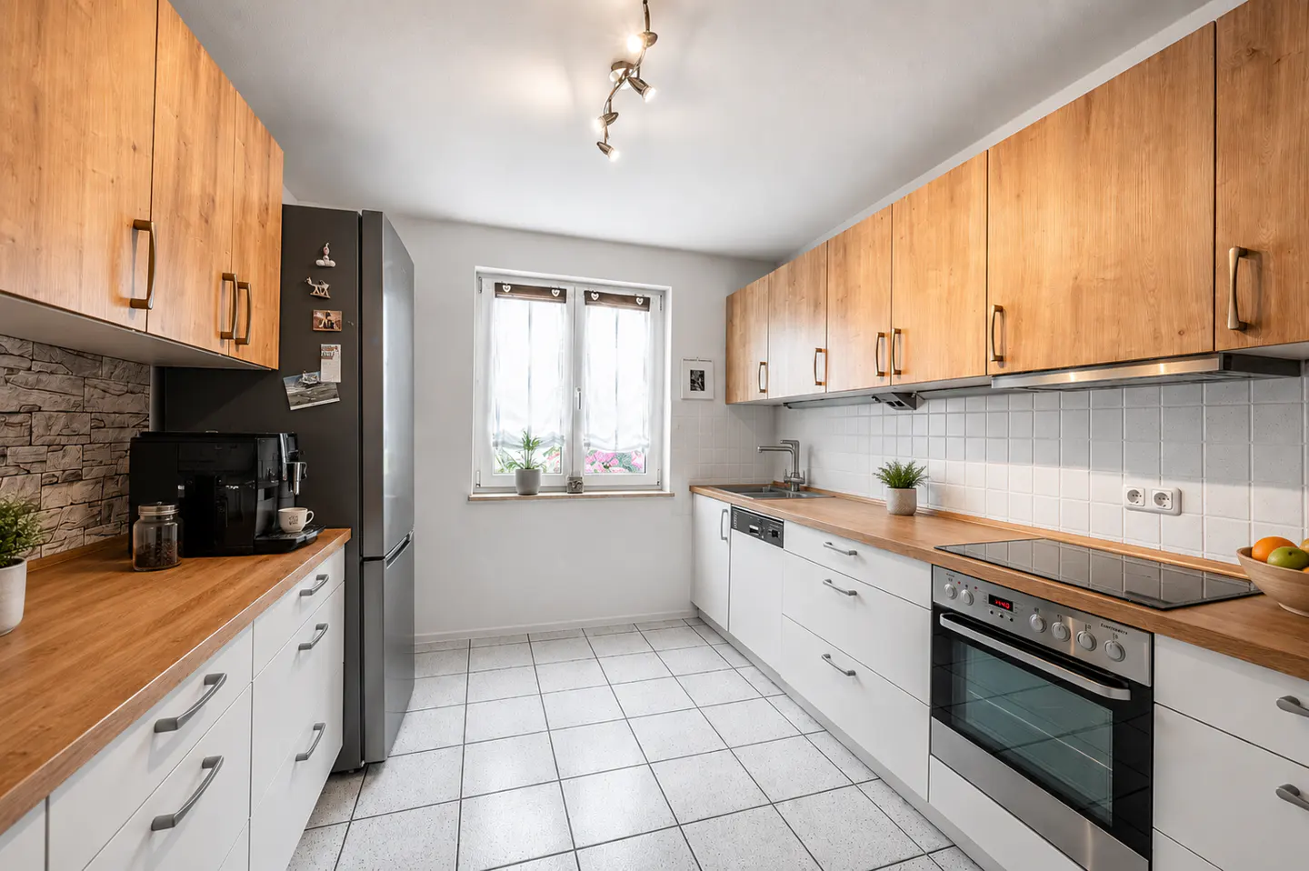 Bright kitchen with white cabinets, wood countertops, and stainless steel appliances. White tile floor and backsplash. Window with sheer curtains.