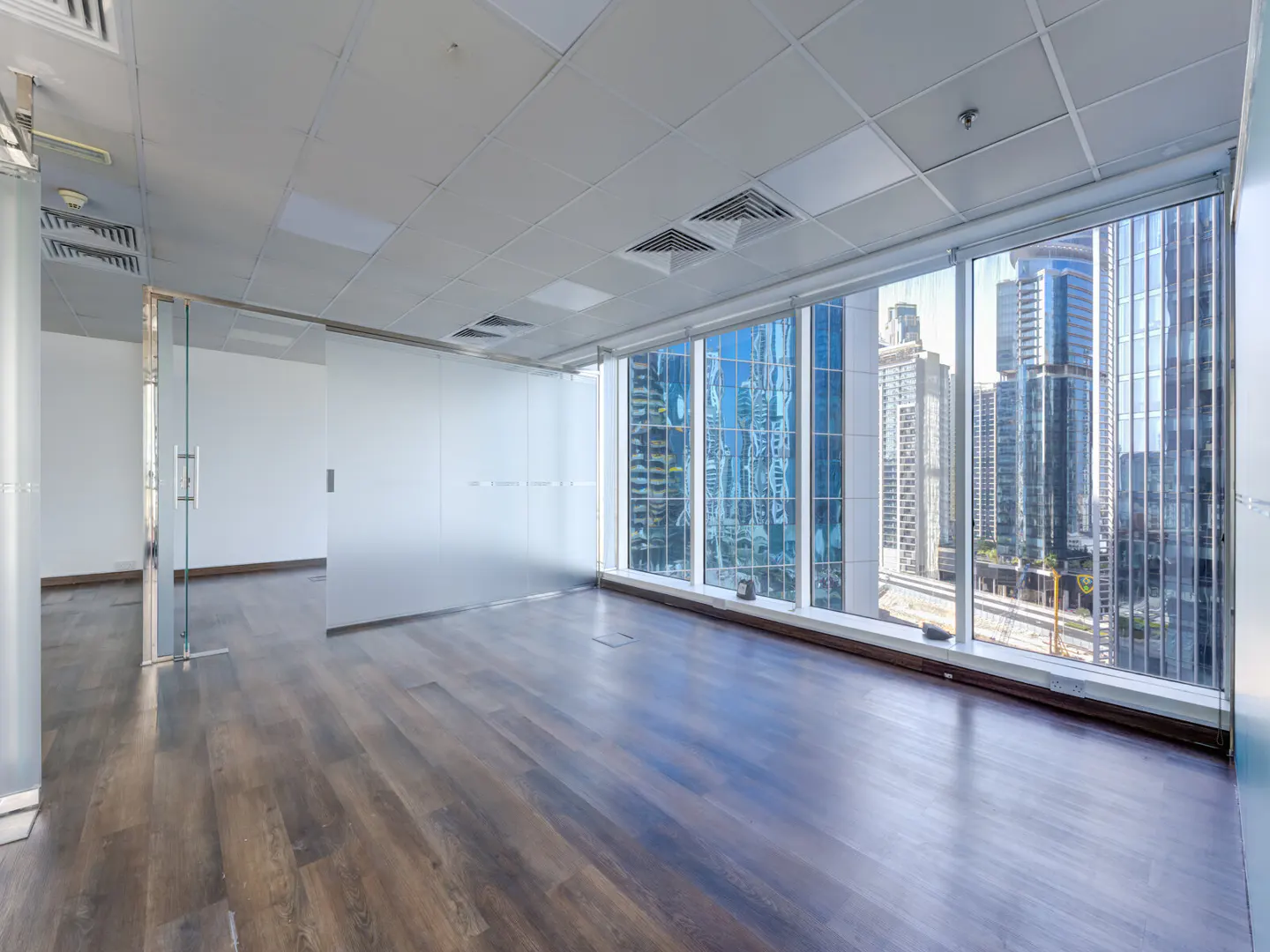 Empty office space with dark wood floors, glass walls, and a large window showing a city skyline.