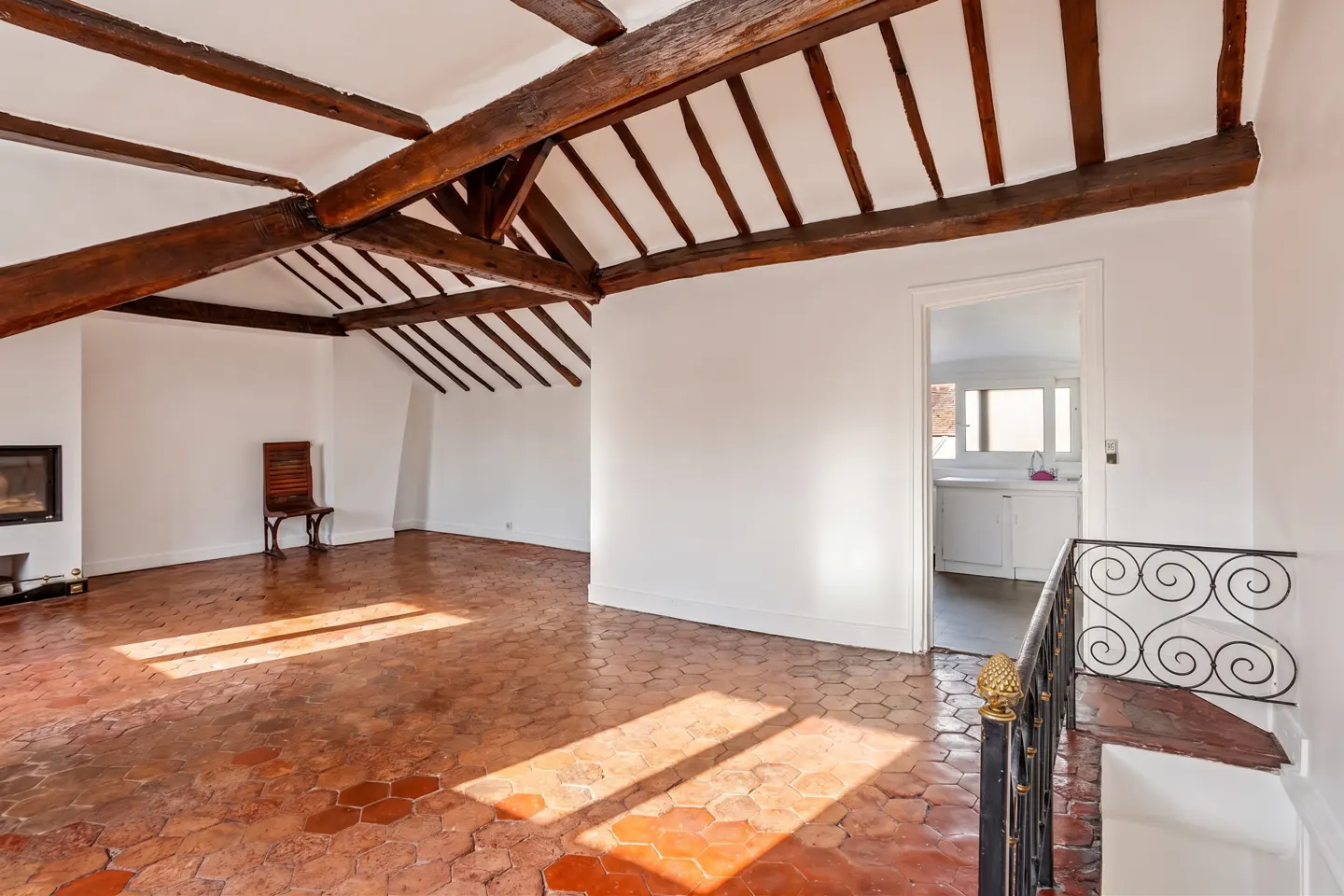 Attic room with white walls, exposed wooden beams, terracotta hexagonal tile floor, a fireplace, and a doorway to a kitchen.