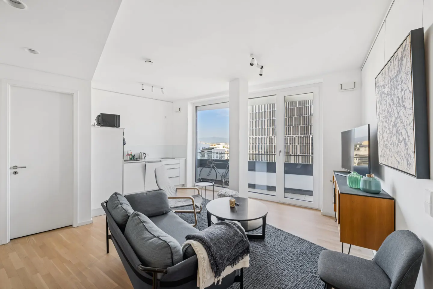 Bright, modern apartment living room with gray sofa, wood floors, and balcony view. White walls and minimalist decor.