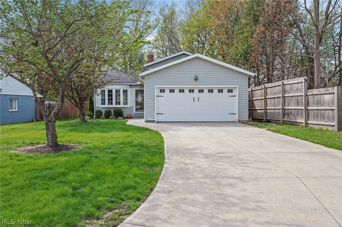 Exterior view of a light gray house with a white garage door and a curved concrete driveway. A green lawn and trees surround the property.