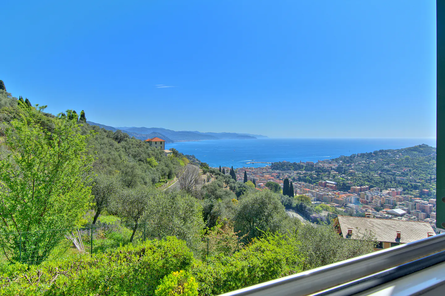 Scenic view of the Italian coastline with green hills, a red-roofed house, and a city by the blue sea under a clear blue sky.