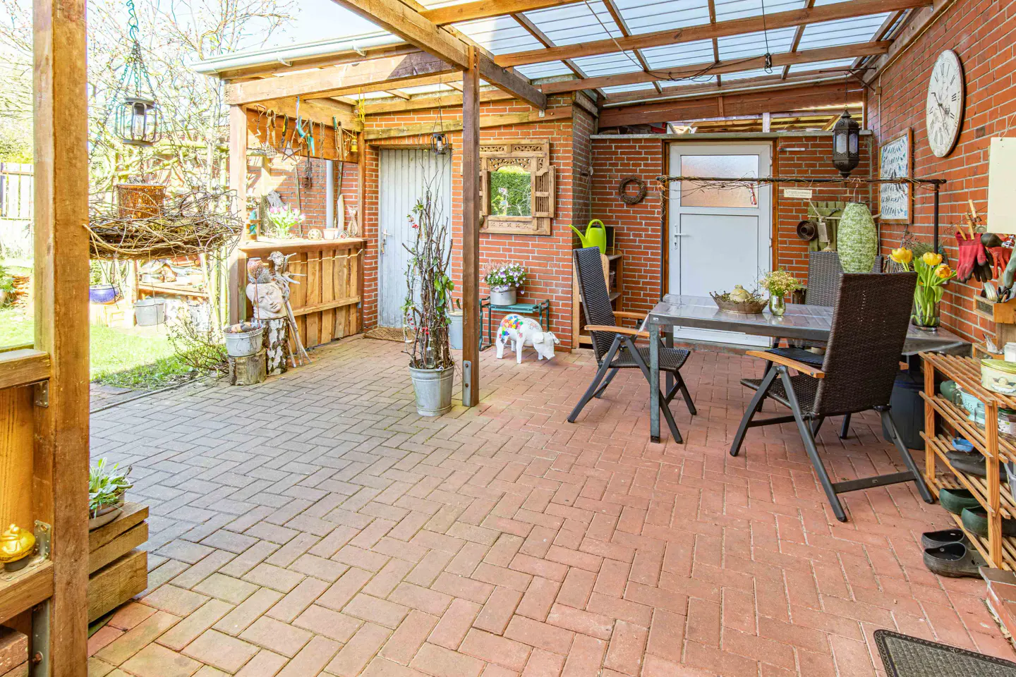 Outdoor patio with brick flooring, a wooden pergola, and a brick wall. A table and chairs sit on the patio, along with plants and decorative items.
