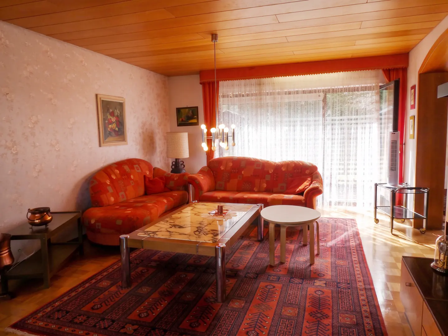 Living room with two orange sofas, a patterned rug, a wooden ceiling, and a large window with sheer curtains.