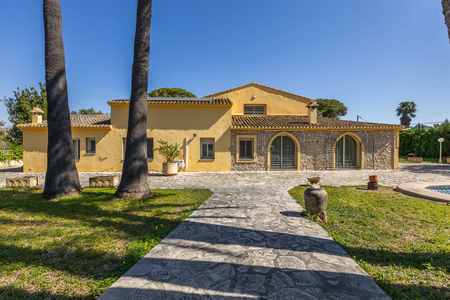Exterior view of a yellow house with stone accents, a tiled roof, and a stone walkway leading to the entrance. Palm trees frame the view.