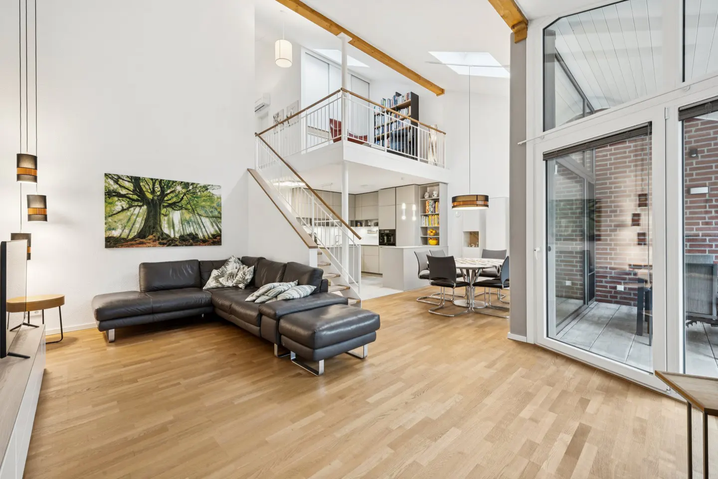 Bright, modern living room with hardwood floors, a black leather sectional, and a staircase leading to a loft overlooking the kitchen. Large windows provide natural light.