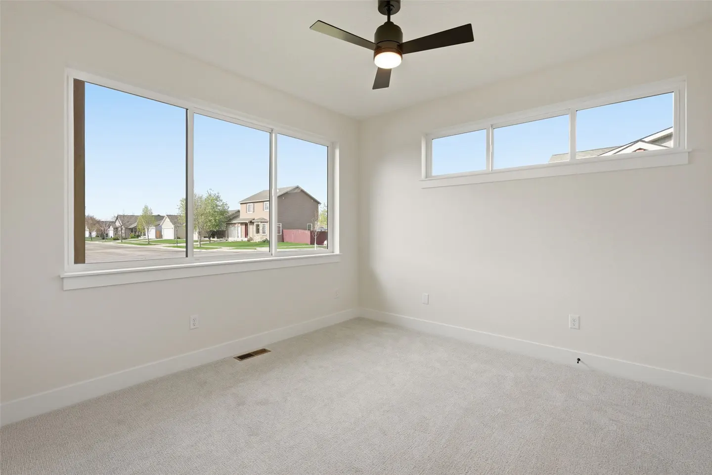 Bright, empty bedroom with gray carpet, white walls, and a ceiling fan. Large windows offer a view of a suburban street.