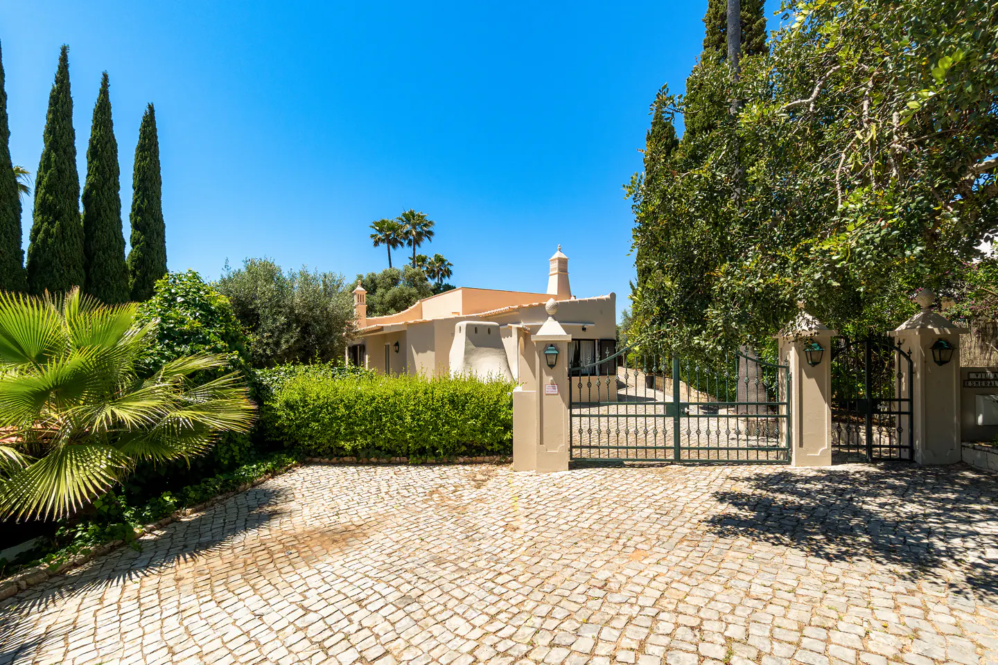 Exterior view of a tan house with a green metal gate and a cobblestone driveway. Tall trees and blue sky in the background.