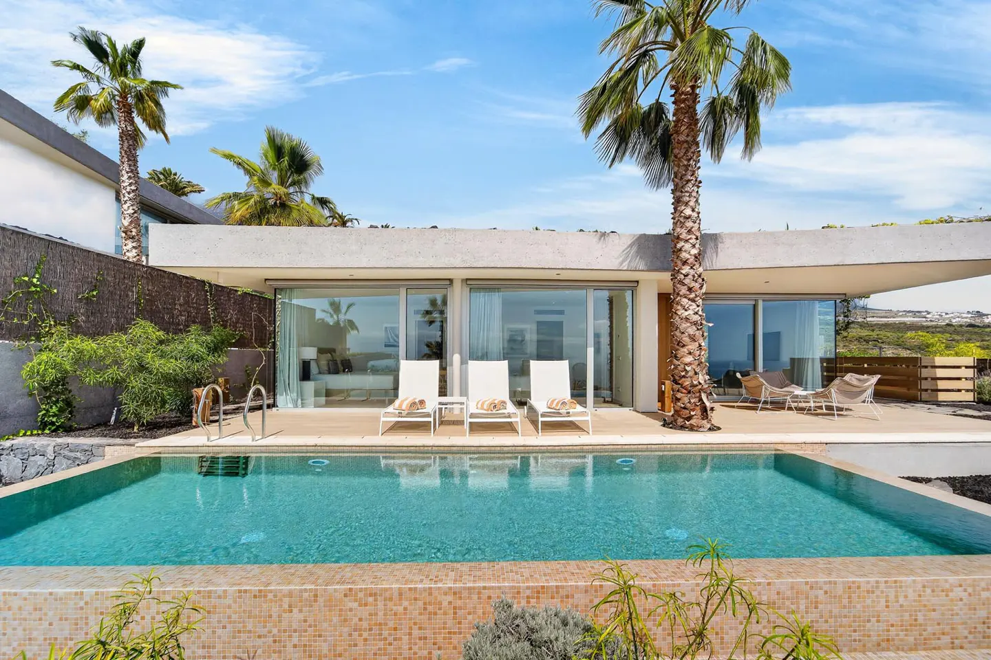 Modern villa with a turquoise pool, palm trees, and white lounge chairs under a blue sky. Large glass doors reflect the ocean view.