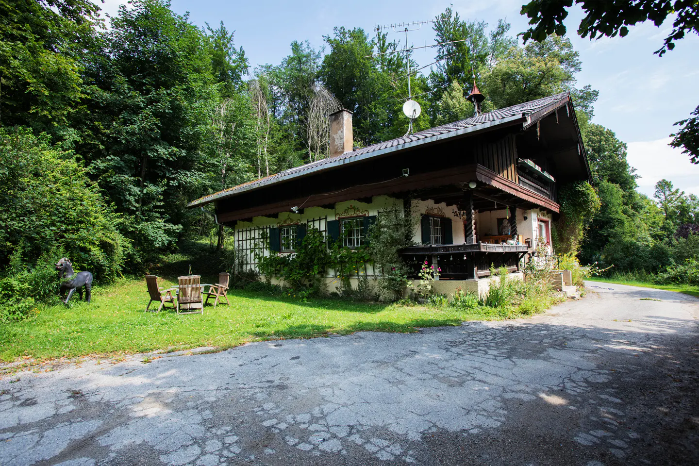 A quaint white cottage with dark brown trim, green shutters, and a stone chimney, nestled among lush green trees. A small patio with chairs is visible.