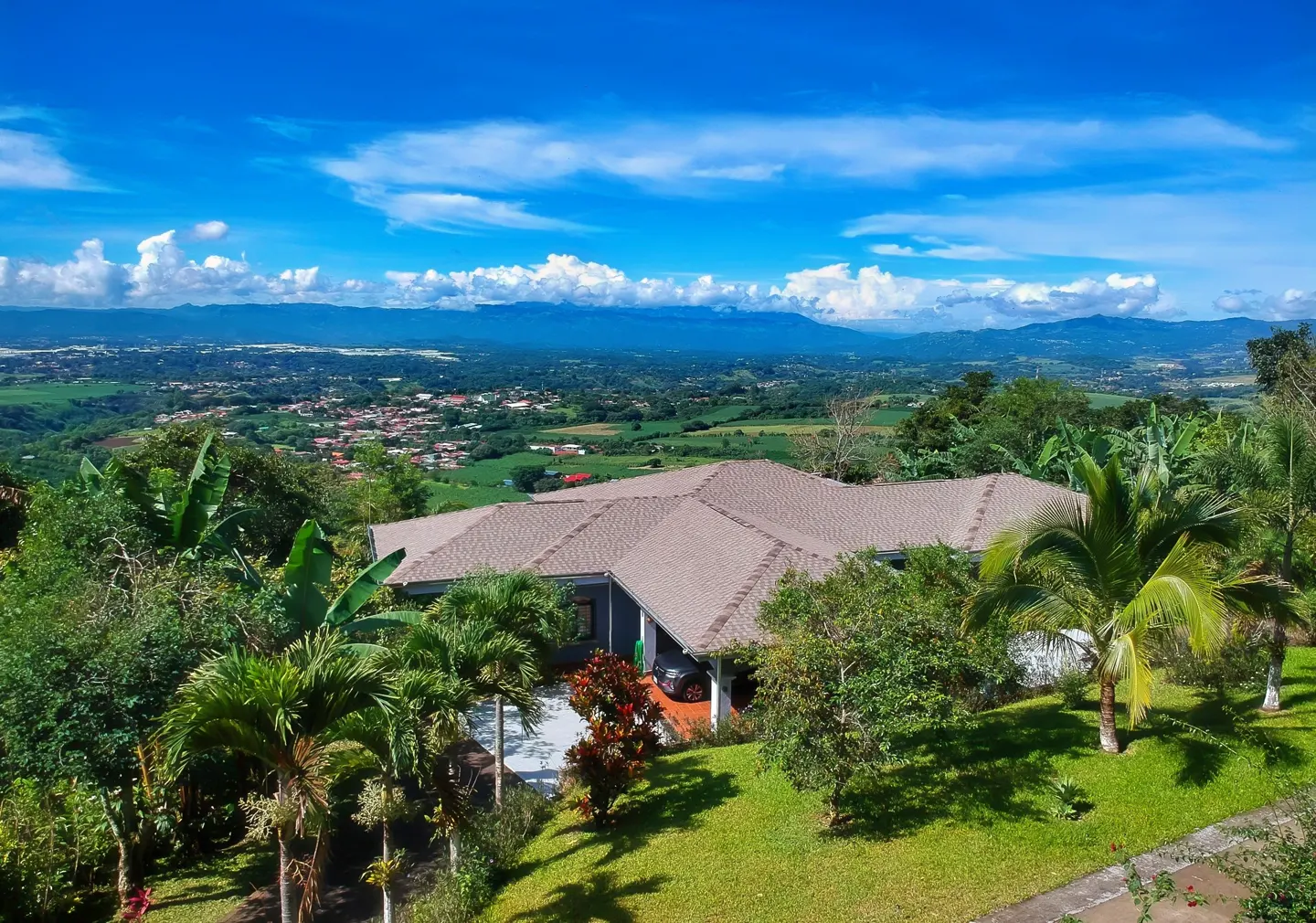 Aerial view of a tan-roofed house surrounded by lush greenery, with a mountain range and blue sky in the background.