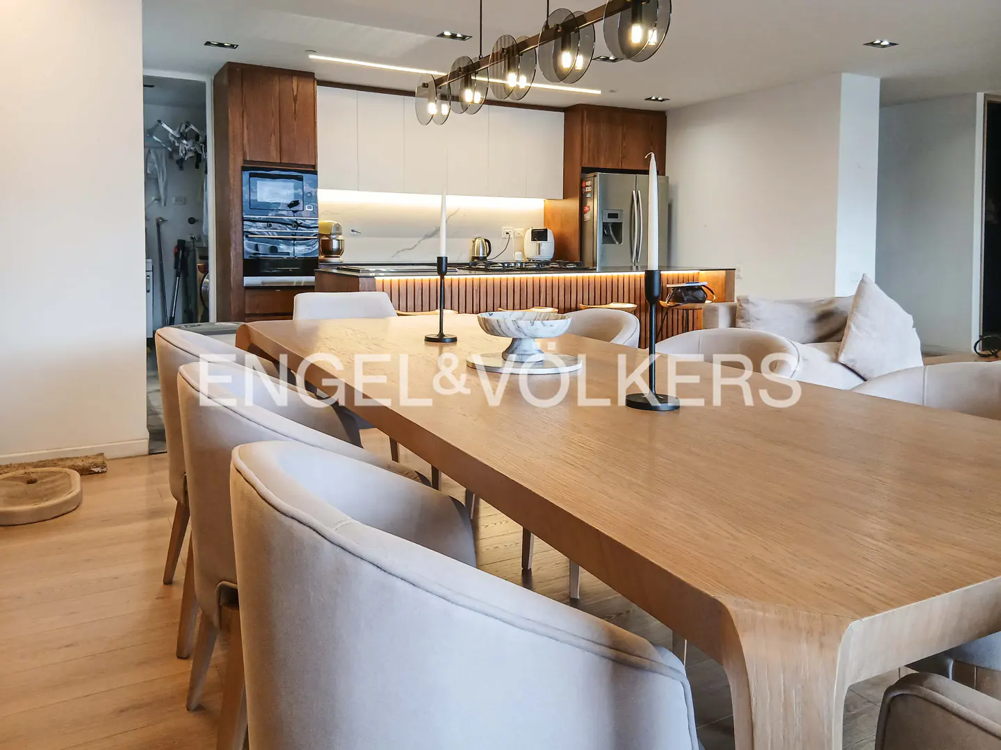 Open-concept dining and kitchen area with a long wooden table and beige chairs. Modern lighting fixture hangs above the table.