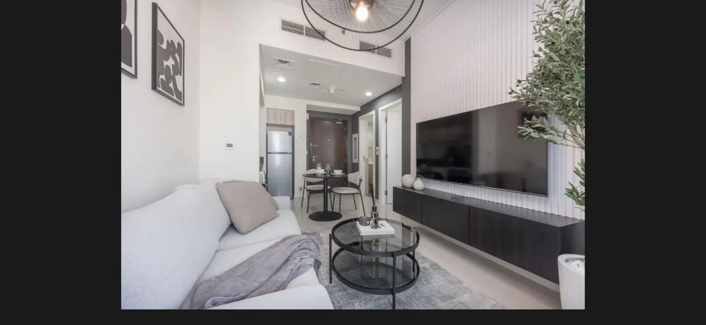 A modern apartment interior with a white sofa, glass coffee table, and a wall-mounted TV. A dining area is visible in the background.