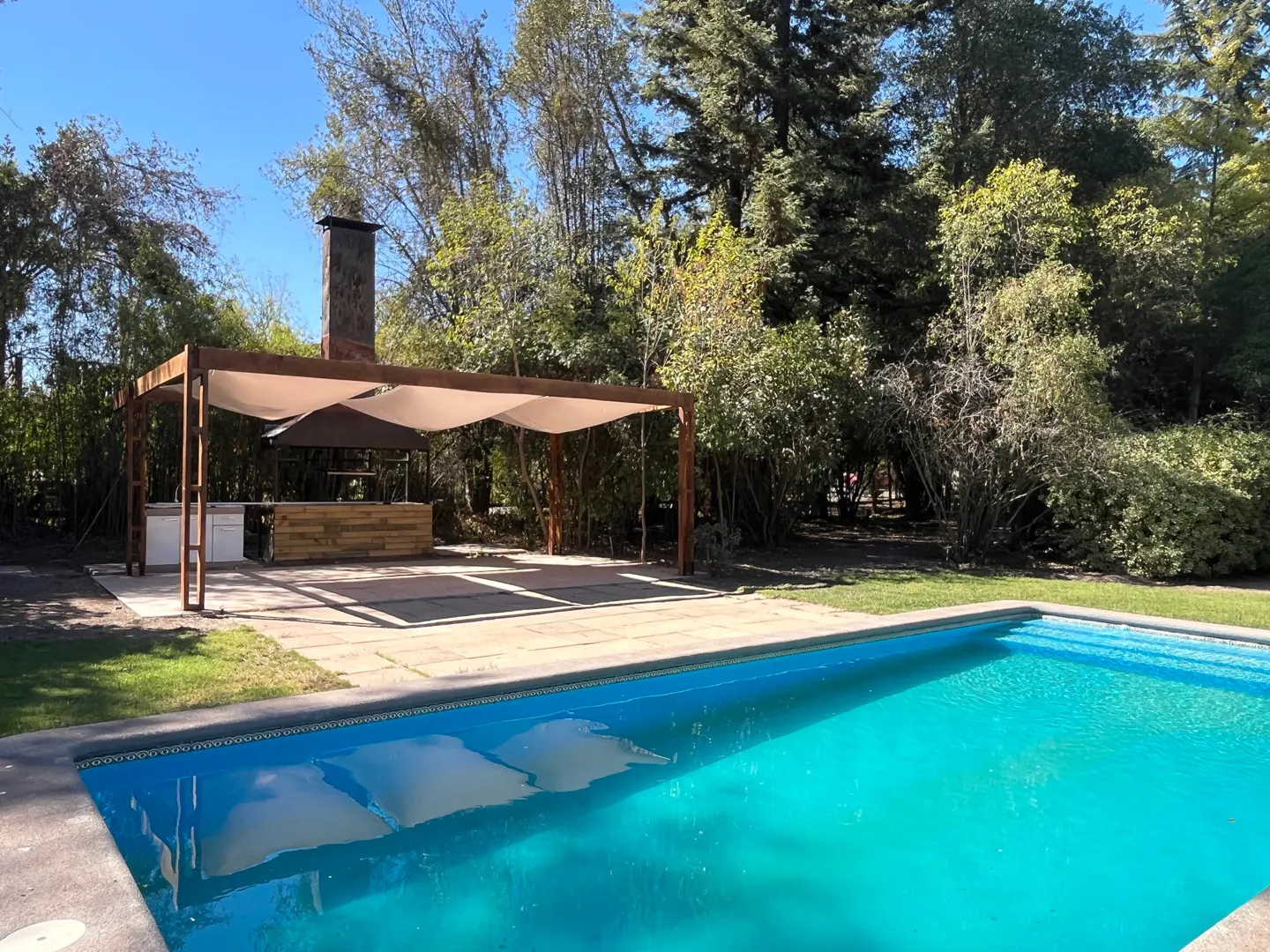 Outdoor pool with turquoise water, stone patio, and covered outdoor kitchen with brick chimney, surrounded by green trees.