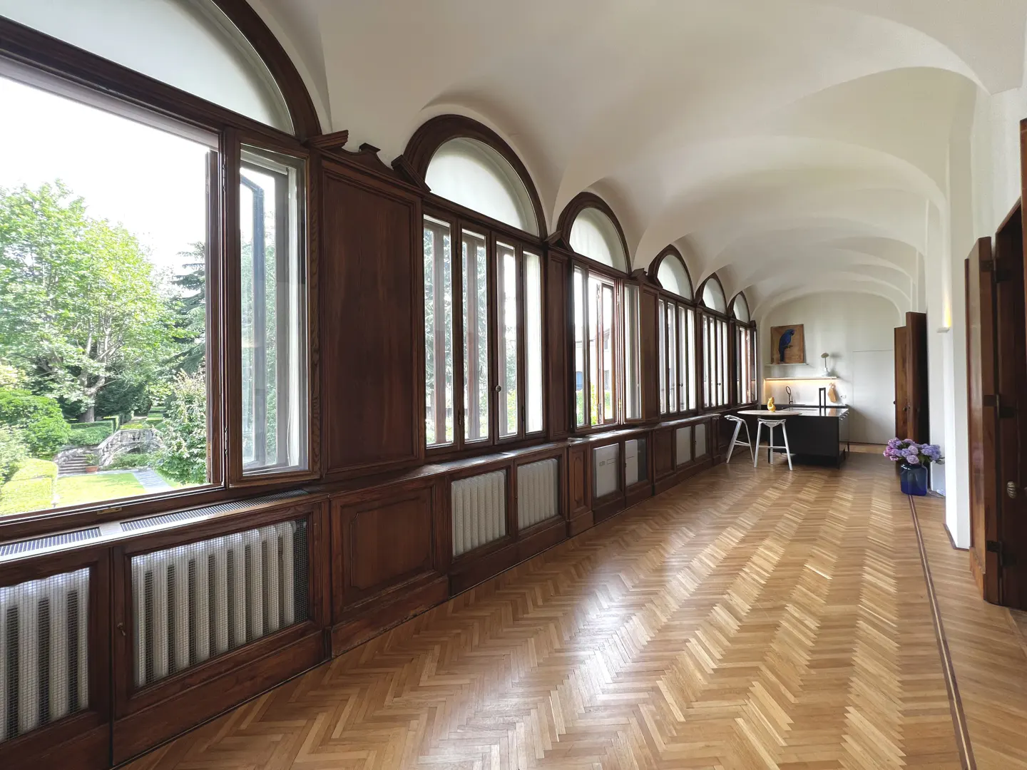 Long hallway with parquet floors, arched windows overlooking a green garden, and a small kitchen area at the end.