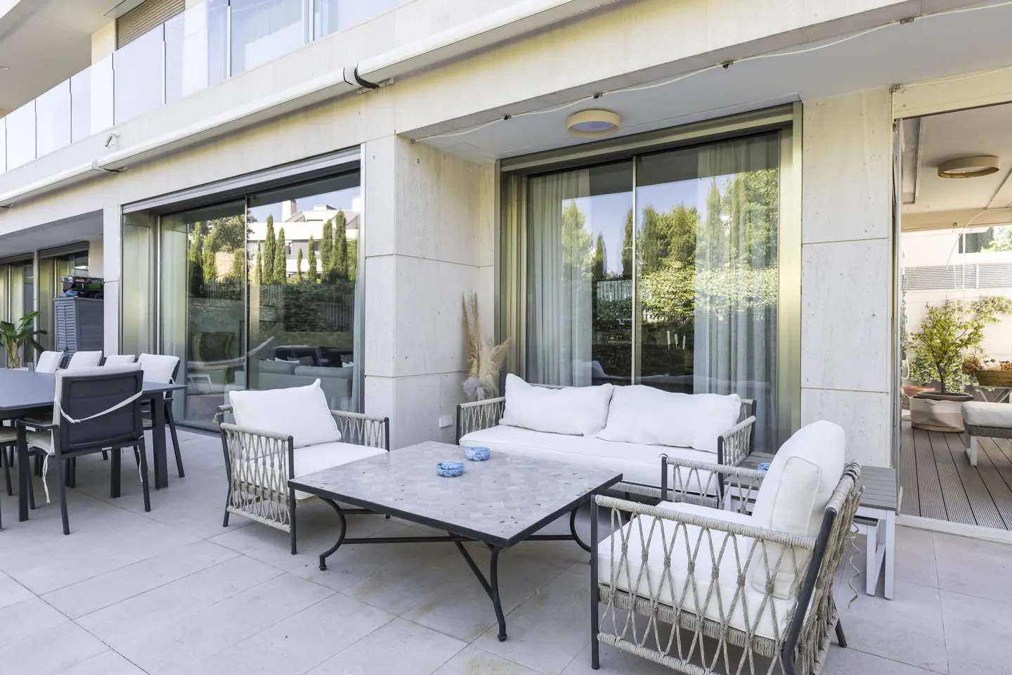 Outdoor patio with white cushioned seating, a dark table, and sliding glass doors leading to a garden view.