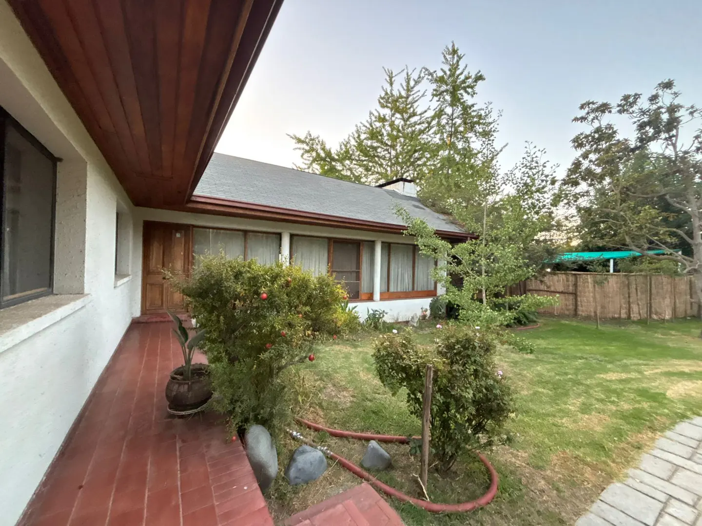 Exterior view of a single-story house with a red brick porch, green lawn, and trees. The house has a gray roof and white walls.