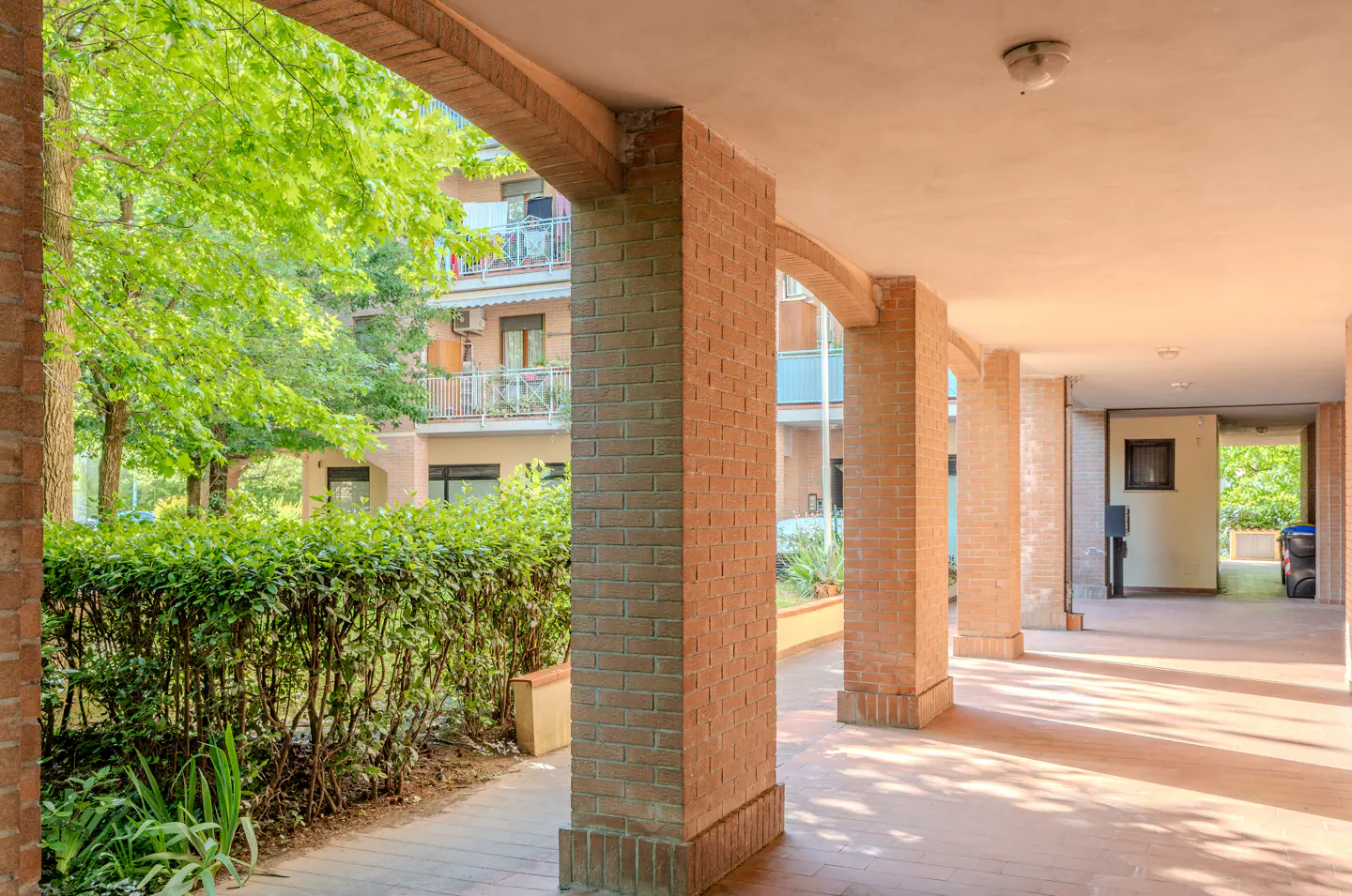 Covered walkway with brick arches and columns, leading to a courtyard with green trees and a building in the background.