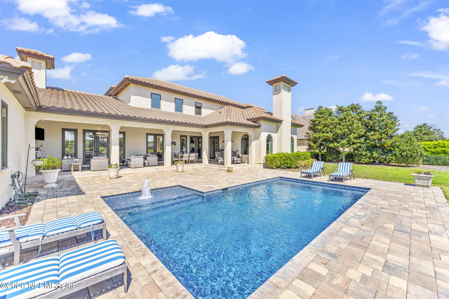 Luxury home exterior with a blue pool, fountain, and striped lounge chairs on a brick patio under a sunny sky.