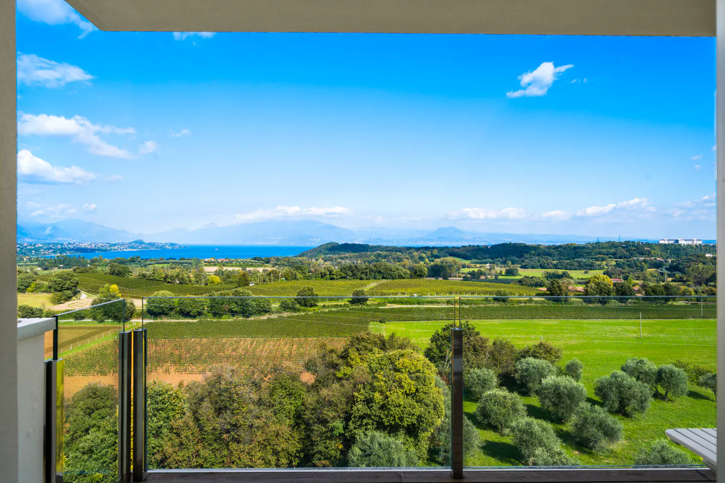 View from a balcony overlooking green fields, trees, and a blue lake under a bright blue sky.