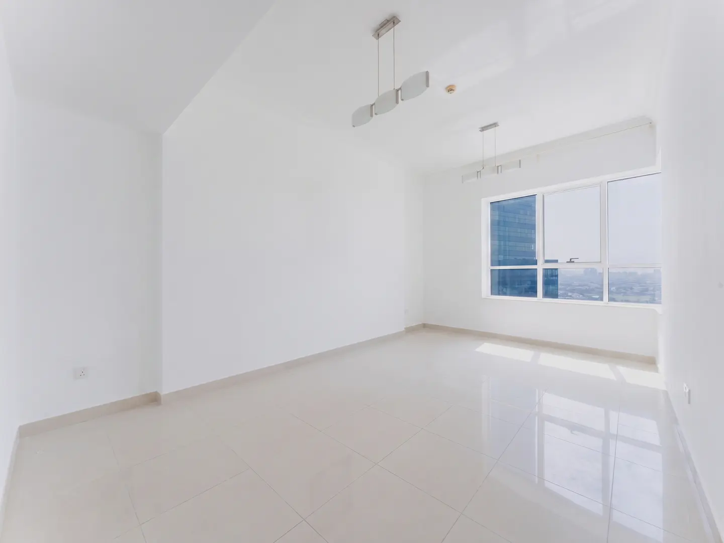 Bright, empty room with white walls, tile floor, and a window showing a city view. Two modern light fixtures hang from the ceiling.