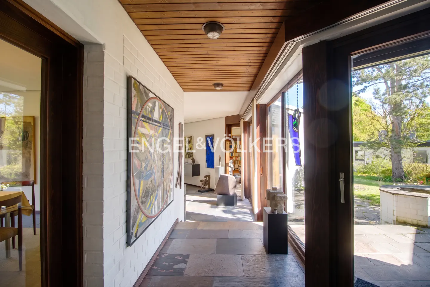 Hallway with stone floors, white brick walls, and wood ceiling. Art hangs on the walls. Engel & Volkers logo is visible. Outside view of trees.