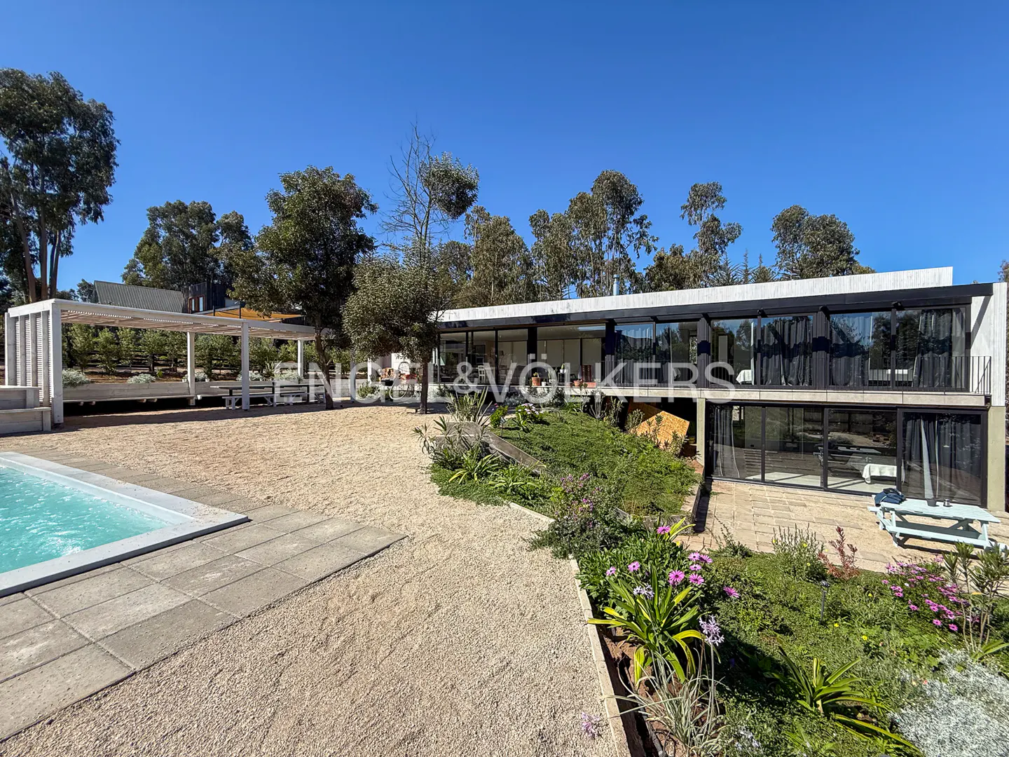Modern two-story house with large windows, a pool, and a white pergola on a sunny day. Trees in the background.