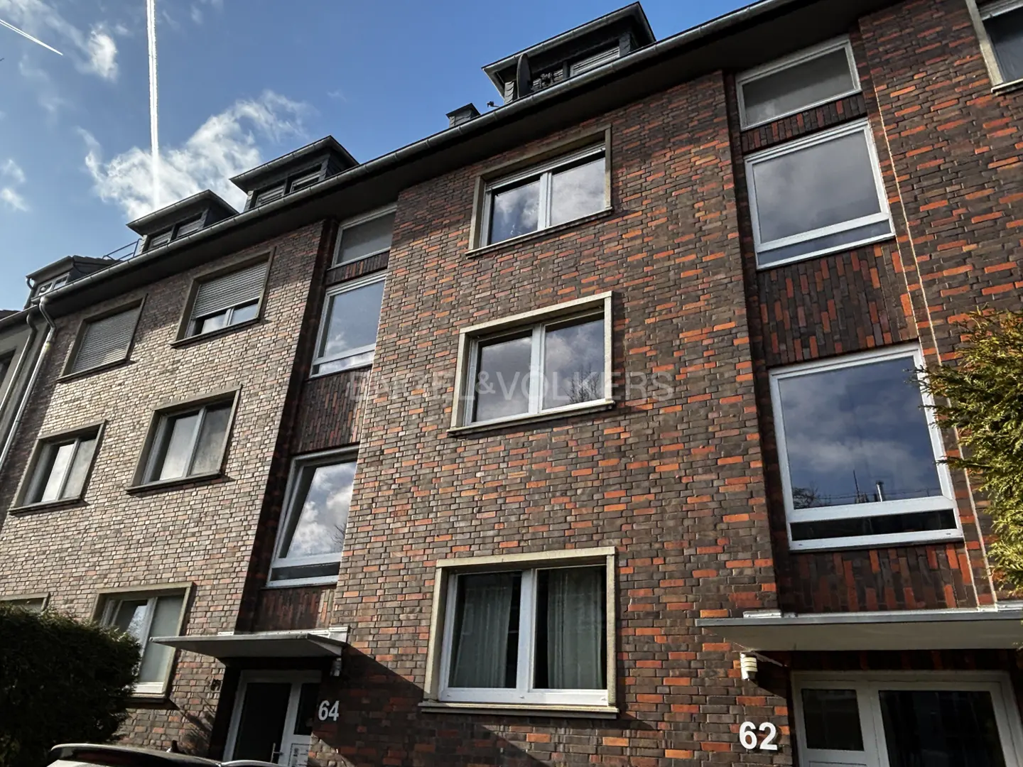 A low-angle shot of a brick apartment building with white-framed windows under a blue sky. The building has unit numbers 62 and 64.