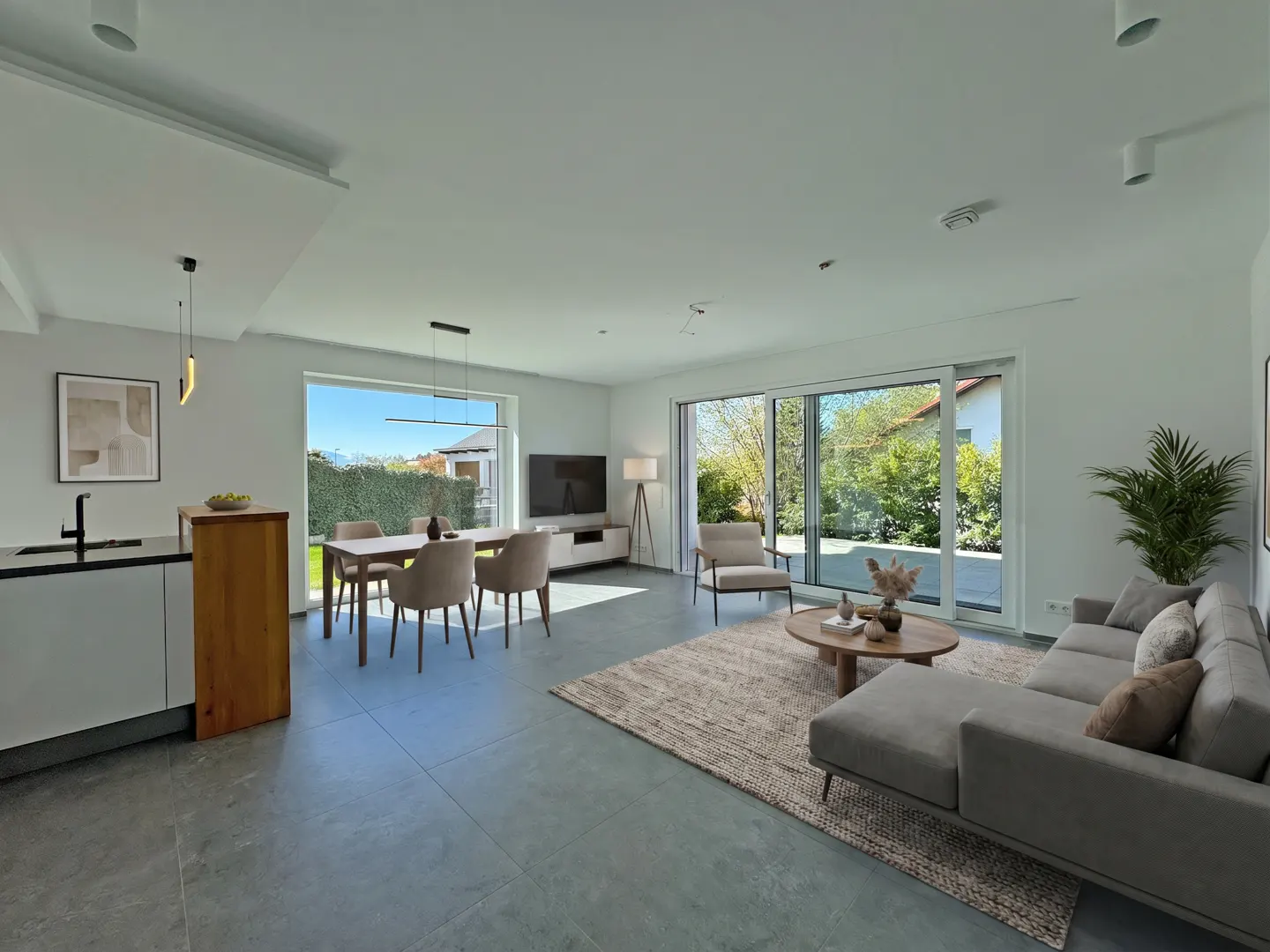 Bright, open-concept living space with gray sofa, dining table, and kitchen island. Large windows offer views of the green outdoors.
