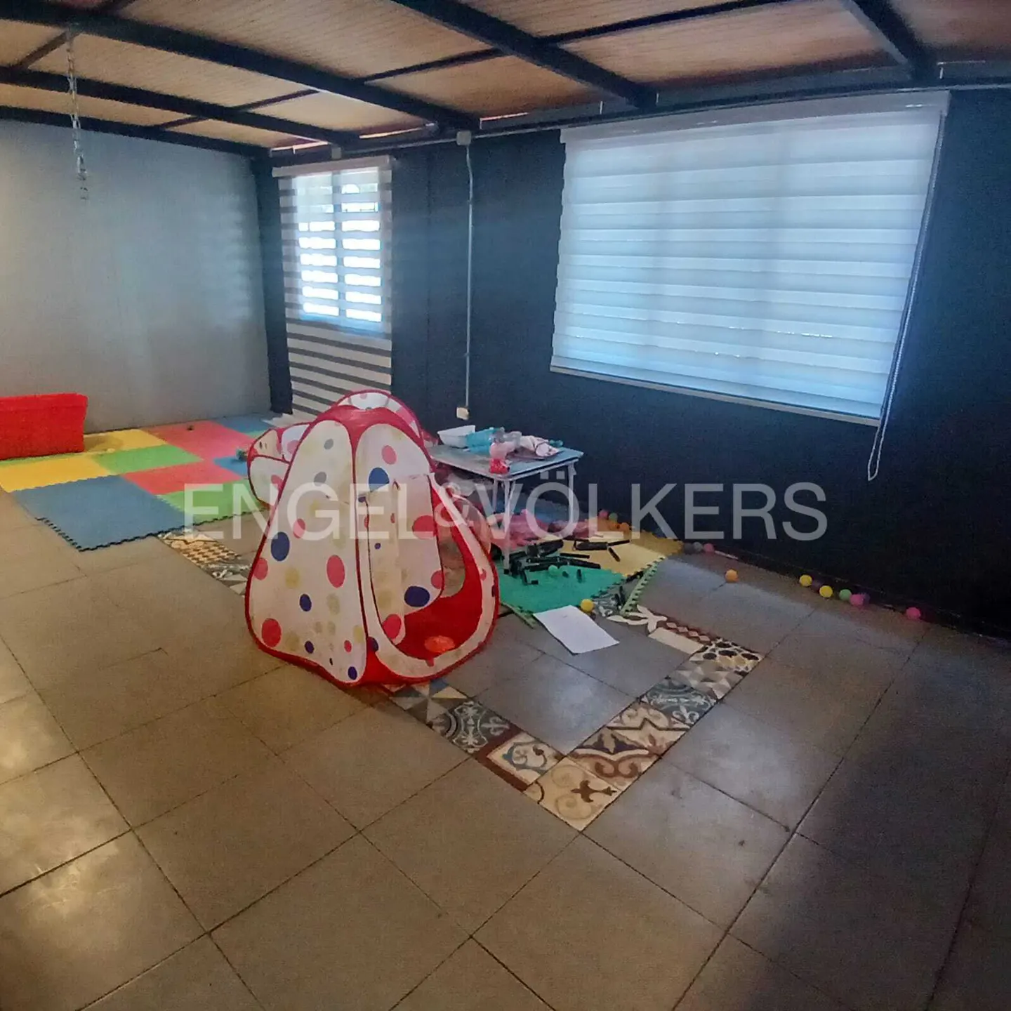 A playroom with a polka-dot tent, colorful floor mats, and a small table with toys. Windows with blinds provide natural light.